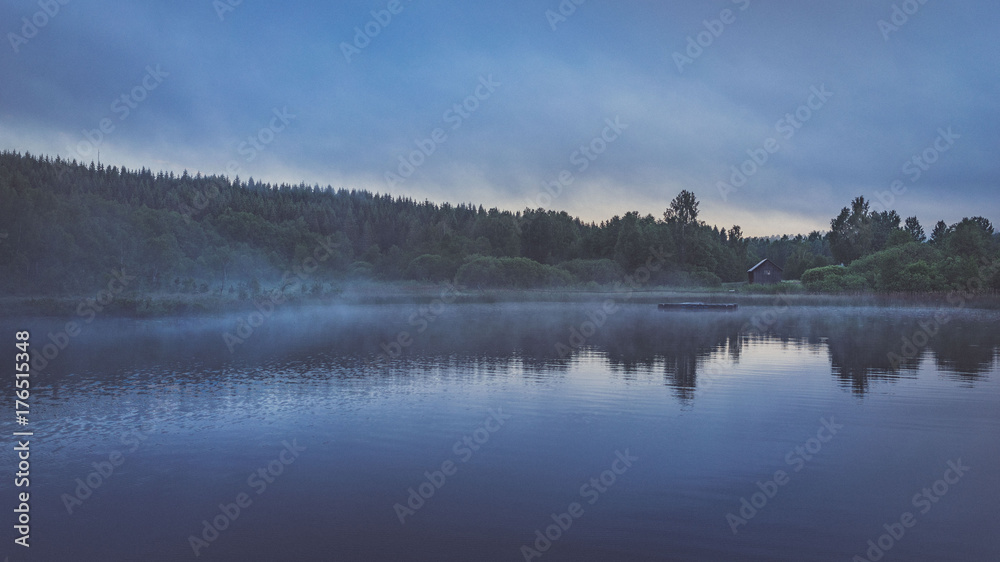 Nebel auf Wasser am Morgen oder Abend vor Ufer mit Bäumen und Wald hinter kleinem Haus in düsterer Stimmung Nebel auf Wasser am Morgen oder Abend vor Ufer mit Bäumen und Wald hinter kleinem Haus in düsterer Stimmung