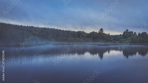 Nebel auf Wasser am Morgen oder Abend vor Ufer mit Bäumen und Wald hinter kleinem Haus in düsterer Stimmung