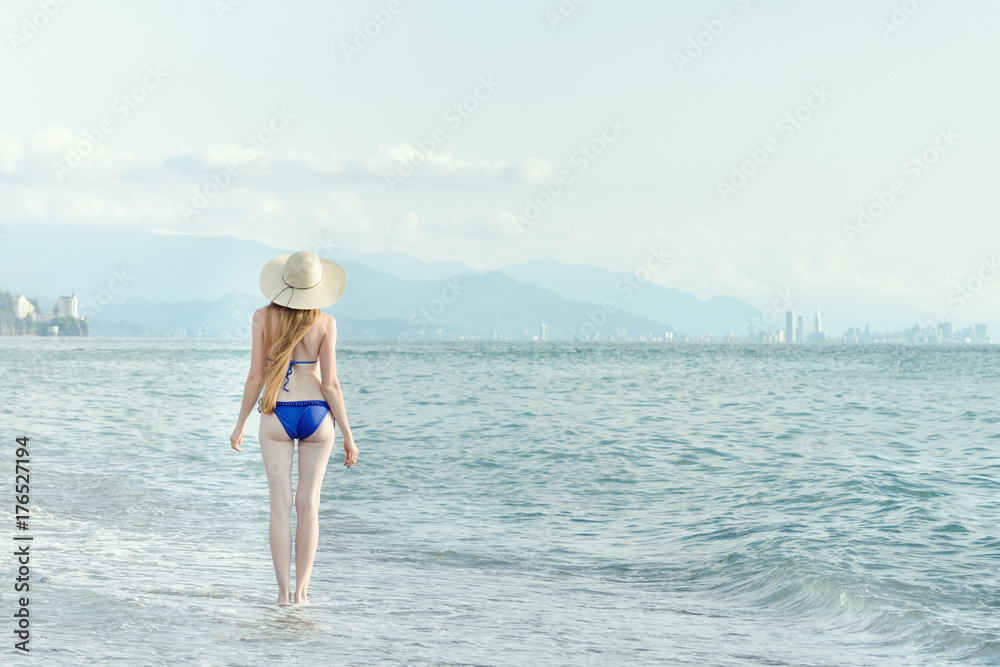 Girl in a bathing suit and hat walks by sea. View from the back