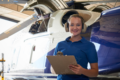 Portrait Of Female Aero Engineer With Clipboard Carrying Out Check On Helicopter In Hangar