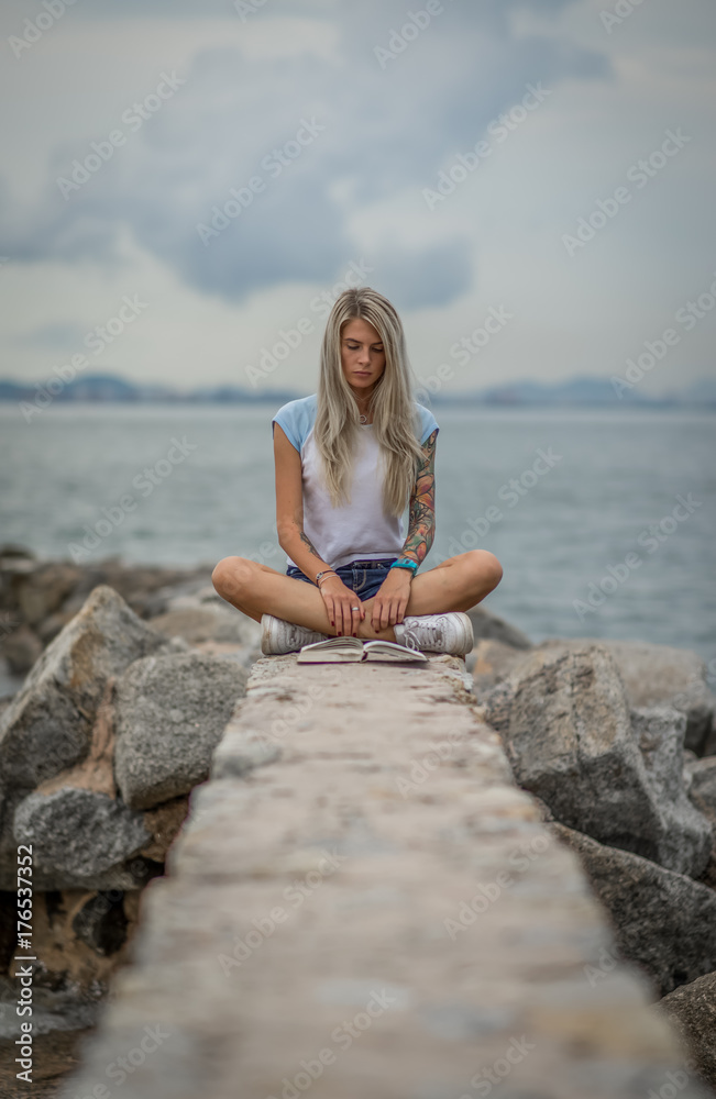 Beautiful blonde woman sitting on a stone embankment and reads book. Hands tattooed. modern, trendy, student Beautiful blonde woman sitting on a stone embankment and reads book. Hands tattooed. modern, trendy, student