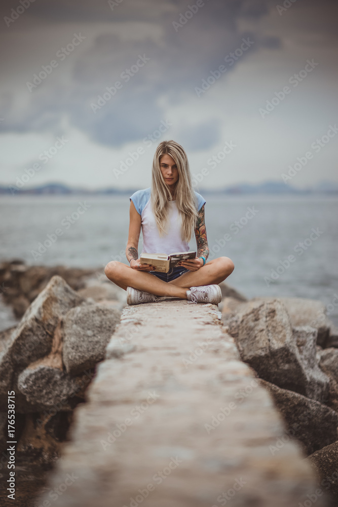 Beautiful blonde woman sitting on a stone embankment and reads book. Hands tattooed. modern, trendy, student