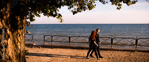 Man and pregnant woman walk through quay on a green tree and blue sea background wide photo