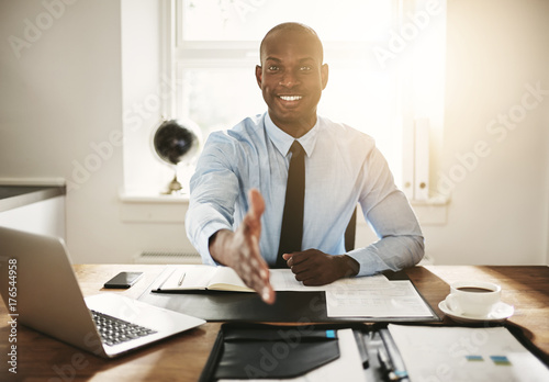 Smiling young executive sitting at his desk extending a handshak