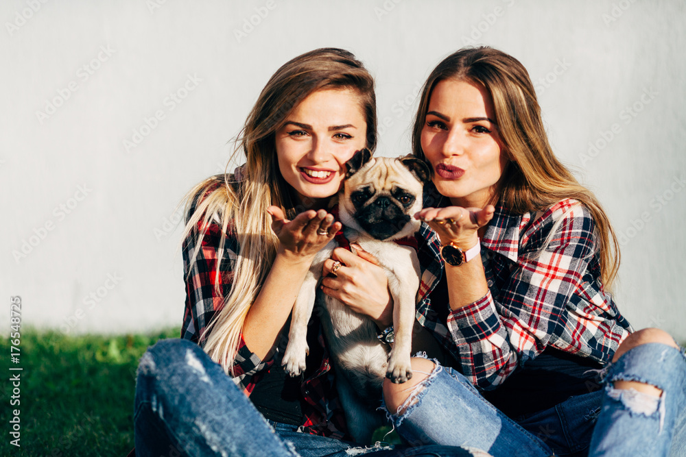 Two beautiful smiling sisters twins and dog pug sitting and sending an air kiss on green grass in the summer park Two beautiful smiling sisters twins and dog pug sitting and sending an air kiss on green grass in the summer park
