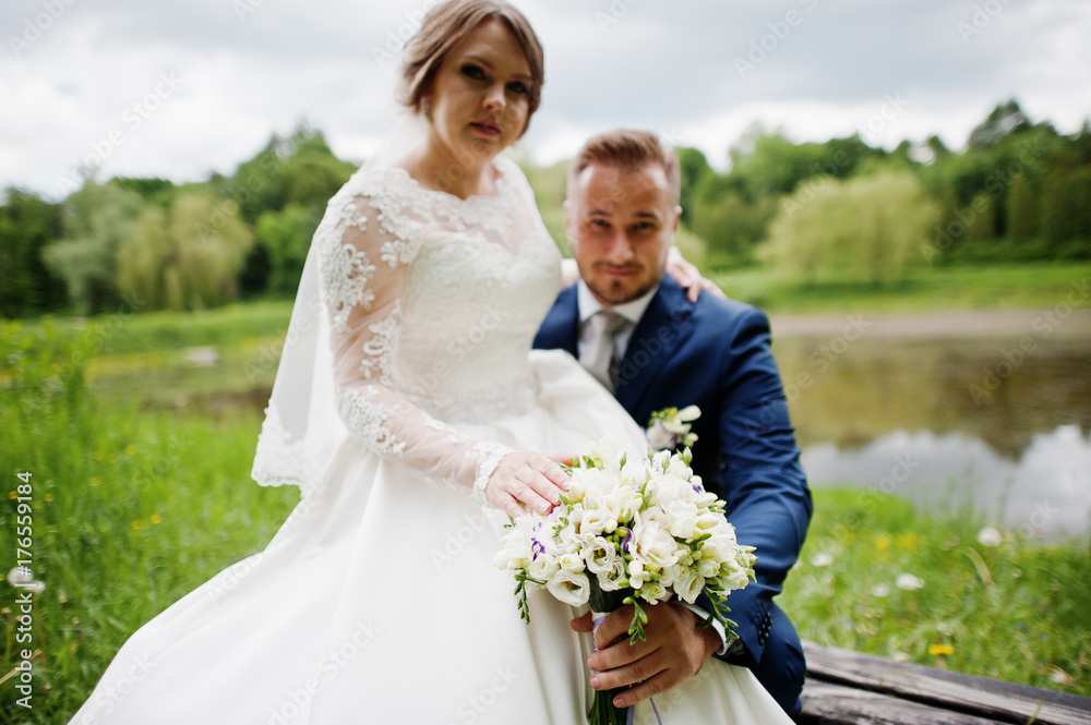 Fantastic bride sitting on a groom's lap in the meadow next to the lake on a wedding day.