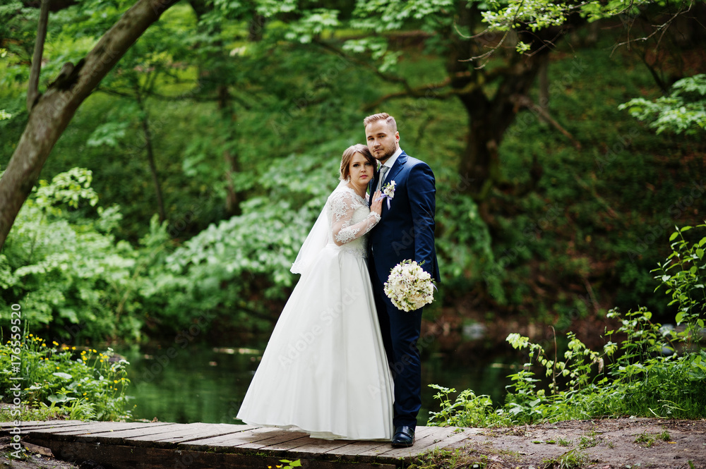 Attractive wedding couple standing and posing on a small wooden bridge next to the pond in the forest.