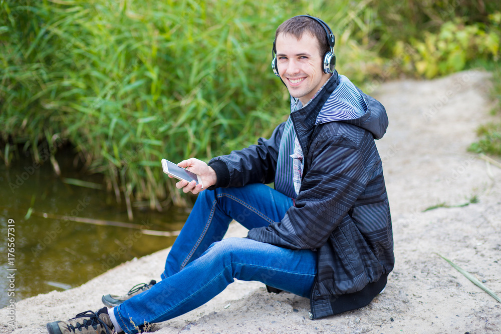 Young man in excellent spirits sits on the river bank with a phone and listens to music Young man in excellent spirits sits on the river bank with a phone and listens to music