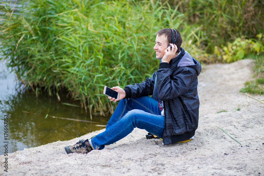 Young man in excellent spirits sits on the river bank with a phone and listens to music Young man in excellent spirits sits on the river bank with a phone and listens to music