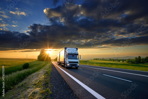 Small truck driving on the asphalt road in rural landscape at sunset with dark clouds