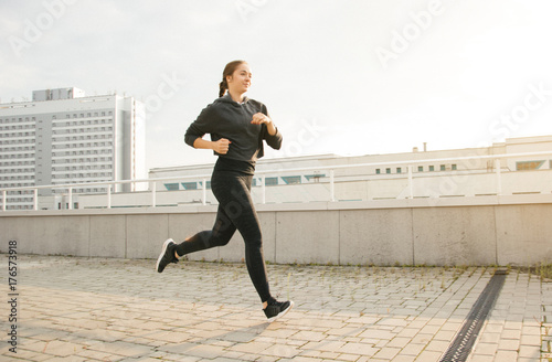 Young attractive lady jogging in the city near buildings. Beautiful happy girl running in the morning outside. Woman trains during the sunrise in the city center before breakfast. 
