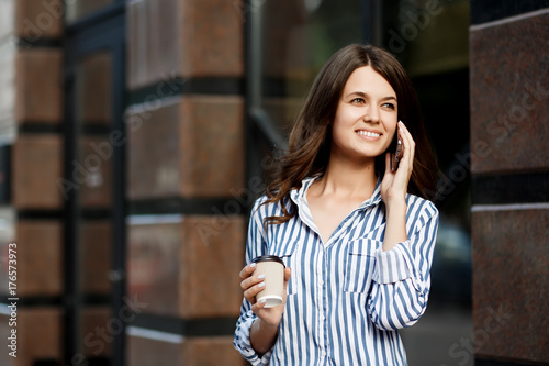 Businesswoman talking on mobile phone, walking along city streets with coffee in hand.