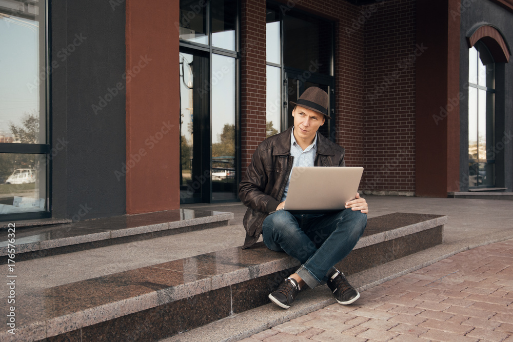 Young handsome boy sitting on a stairs near office building in the center of the city, working on his notebook, using mobile phone and having break outside on a sunny warm autumn day. Happy freelancer