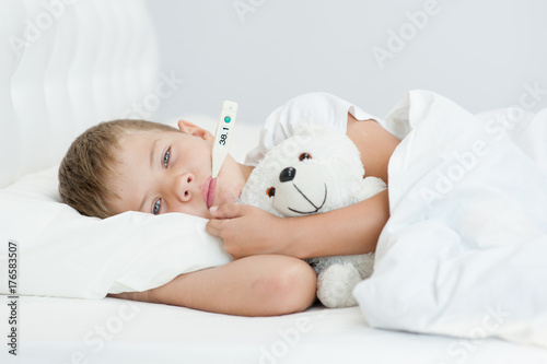 Sick boy with thermometer in mouth lying in bed and hugging a toy bear