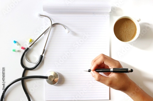 Hand Holding Pen in Flat lay photo with Notebook , Coffee Cup , Stethoscope ,Medical Capsuls on white background
