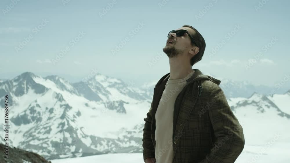 Young bearded caucasian man in brown jacket and sunglasses breathing deeply at snowy mountains with scenic view on sunny day