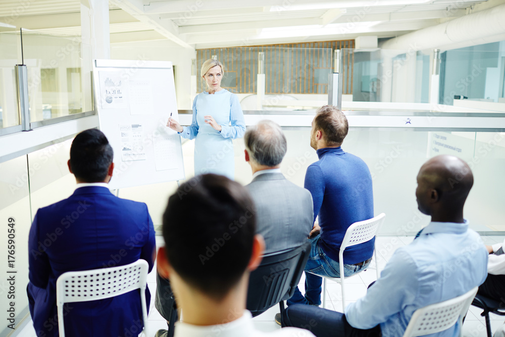 Confident mature speaker making report for audience by whiteboard at seminar or conference Confident mature speaker making report for audience by whiteboard at seminar or conference