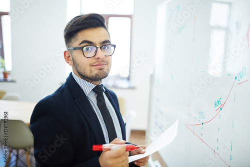 Businessman with red highlighter and paper standing by whiteboard with drawn graph of financial rate