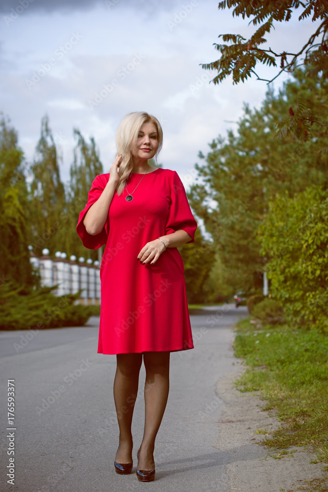 Portrait of pretty middle-aged woman in a red oversize dress in a cottage town, a beautiful blonde hair lady is walking in the open air Portrait of pretty middle-aged woman in a red oversize dress in a cottage town, a beautiful blonde hair lady is walking in the open air