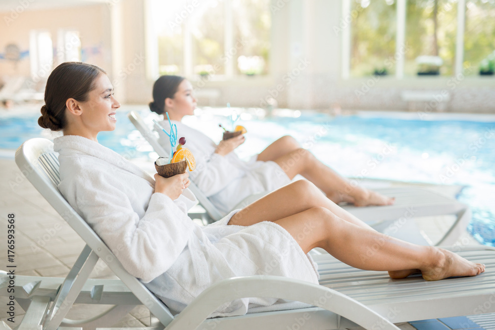 Obraz premium Serene girls in white bathrobes lying on deckchairs by swimming-pool and enjoying pinacolada from coconuts