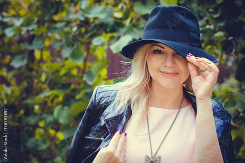 Mature stylish blonde hair woman dressed in leather black jacket and black hat walk in autumn garden with a good mood. Lady with pretty accessorize and casual style