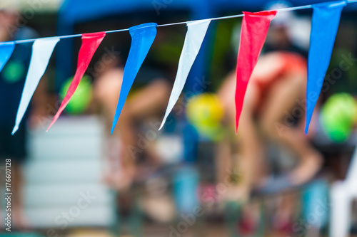 sport multi-color sling flags hanging over swimming pool, with blurred background of swimmers getting ready to start