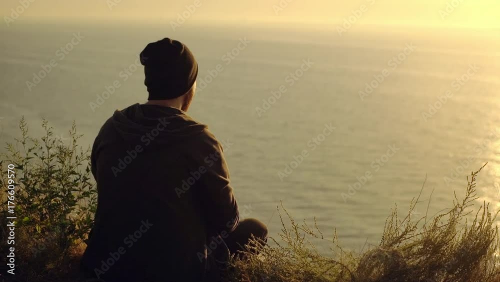 view from back of hipster young guy wearing black clothes sitting on the edge of hill playing with hacky sack throwing it up at warm sunset over sea slow motion