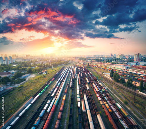 Aerial view of colorful freight trains. Railway station. Wagons with goods on railroad. Cargo trains. Heavy industry. Industrial scene with trains, city buildings and cloudy sky at sunset. Top view 