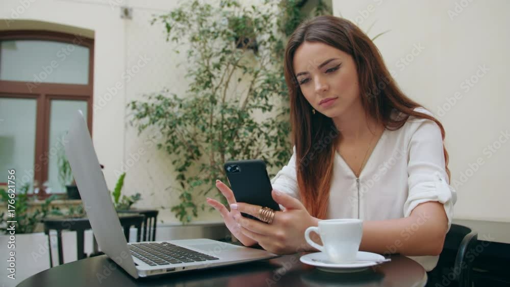 A beautiful girl sitting at the table in the pub and using her laptop and a phone. Medium shot. Soft focus. A beautiful girl sitting at the table in the pub and using her laptop and a phone. Medium shot. Soft focus.