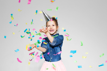  little girl in a denim shirt and a pink tulle from a tulle in a festive cap stands on the white background with confetti and smiles
