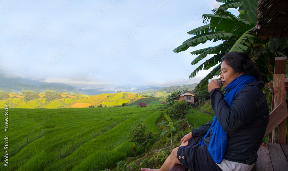 Naklejka premium Woman tourism sitting on wood balcony smelling and drinking hot coffee in morning time and enjoy to see view of green rice field at Napongpeng,Chiangmai-Thailand