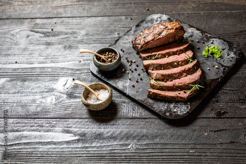 Tableau sur toile Sliced grilled roast beef with salt and pepper on marble plate on wooden old rustic table Black background
