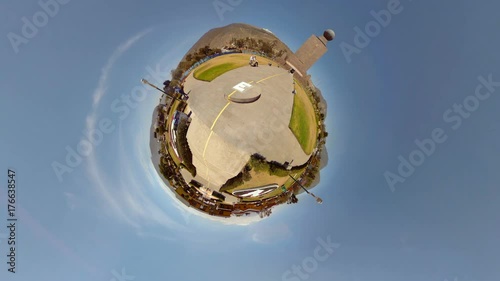 A spherical 360 tiny-planet view of the Ciudad Mitad del Mundo equator monument complex near Quito, Ecuador, stone structures, green lawns and surrounding trees under a clear blue sky.