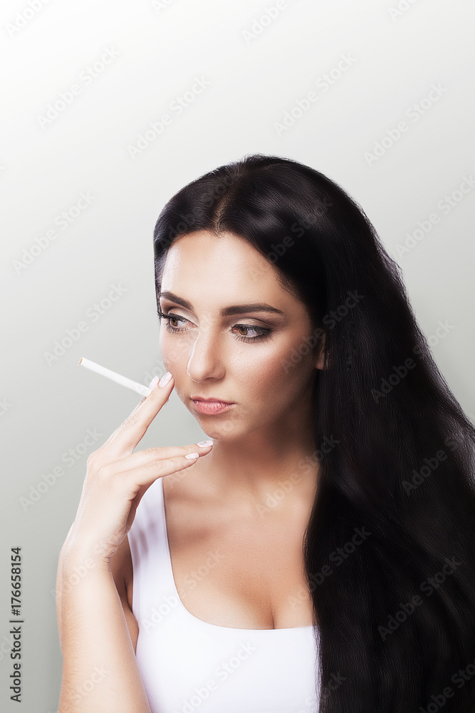 Smoke a young girl Beautiful young woman smoke a cigarette. Beautiful portrait. Girl with long black hair. The concept of health. On a gray background. Smoke a young girl Beautiful young woman smoke a cigarette. Beautiful portrait. Girl with long black hair. The concept of health. On a gray background.