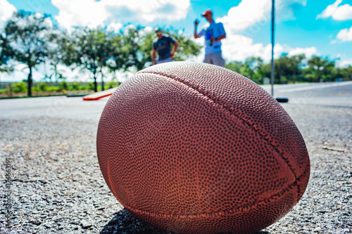 close up of an american football on the ground with people playing cornhole in the background while tailgating before the game