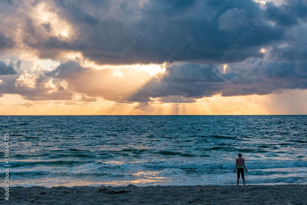 silhouette of female on a sandy beach enjoying the sun rising behind the clouds