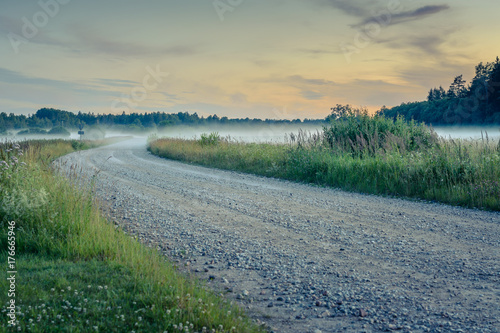  Sunset in countryside at mid summer. Hay rolls and road in misty weather with clear sunny sky. Beautiful nature in Latvia in August. 