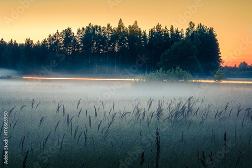  Sunset in countryside at mid summer. Hay rolls and road in misty weather with clear sunny sky. Beautiful nature in Latvia in August. 