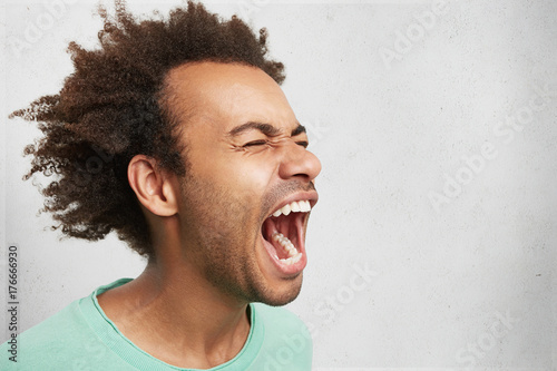 Horizontal portrait of man with dark skin and Afro hairstyle screams in despair, opens mouth widely, being in panic. Frustrated mixed race man poses against white studio background with copy space photo