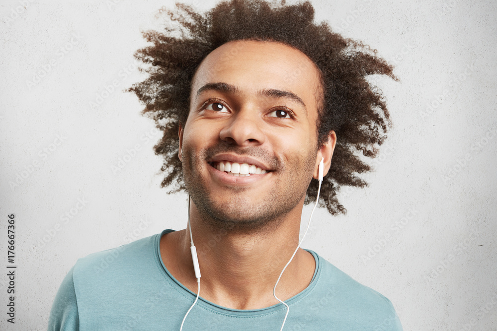 Fotka „Portrait of cool young black man with curly hair, has cheerful  expression, smiles broadly, demonstrates white perfect teeth, listens to  jazz or pop music, looks up dreamy, enjoys good atmosphere“ ze