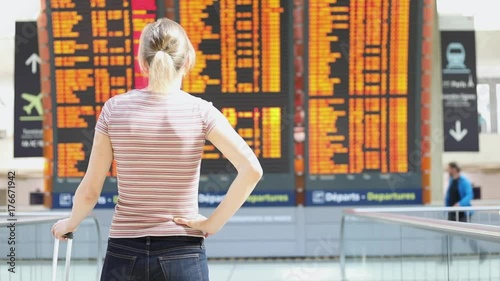Woman traveler with suitcase reading the arrival departure electronic flight information board in the airport and looking at her smartphone before heading to the check-in counter