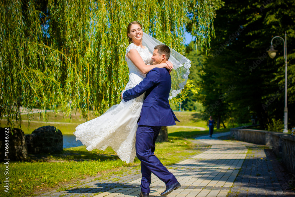 Fototapeta premium Young happy wedding couple dancing and having fun in sunny wedding day. Groom spinning bride. Dress develops in the wind. Groom holding happy emotional bride. The bride is the size of the plus size. 