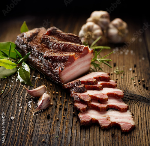 Smoked bacon on a wooden rustic table with addition of fresh aromatic herbs. Natural product from organic farm, produced by traditional methods