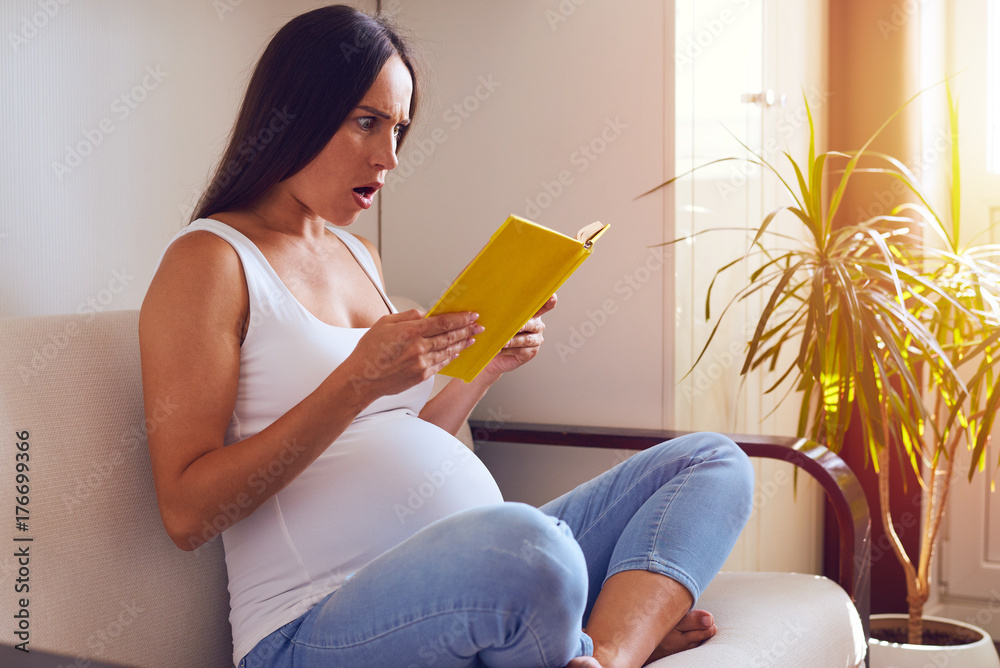 Surprised pregnant woman reading book while sitting on sofa Surprised pregnant woman reading book while sitting on sofa