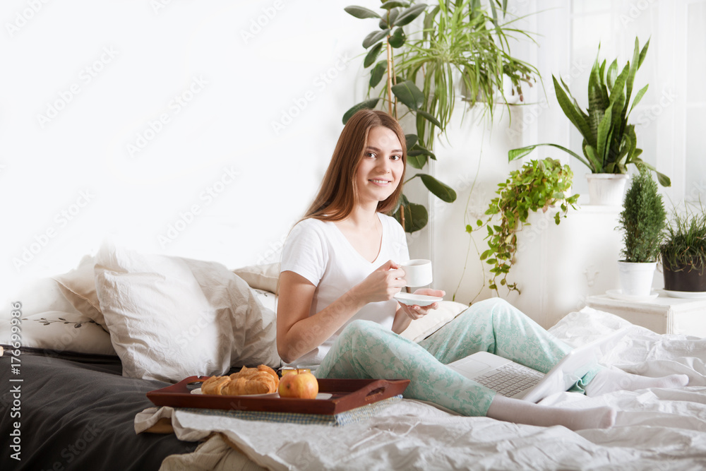 Beautiful young girl working on a laptop sitting on the bed in the house.Breakfast in bed for young beautiful woman Beautiful young girl working on a laptop sitting on the bed in the house.Breakfast in bed for young beautiful woman