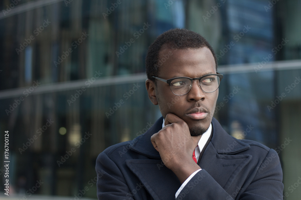 Horizontal headshot of young dark-skinned male standing in coat outdoors looking sideways with expression of suspicion and doubt while finding most effective way out of difficult situation in business Horizontal headshot of young dark-skinned male standing in coat outdoors looking sideways with expression of suspicion and doubt while finding most effective way out of difficult situation in business