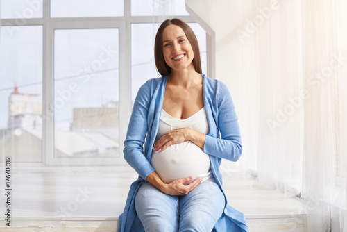 Affectionate smiling woman holding belly sitting against window