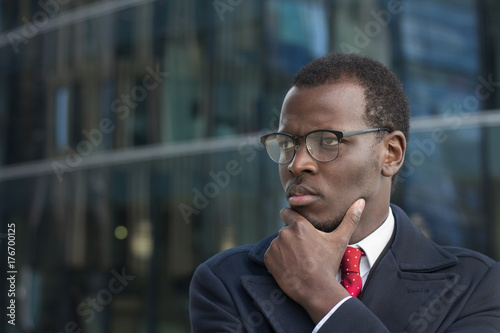Horizontal headshot of attentive African american executive standing outdoors, casting side look through eyeglasses scratching chin with fingers, thinking photo