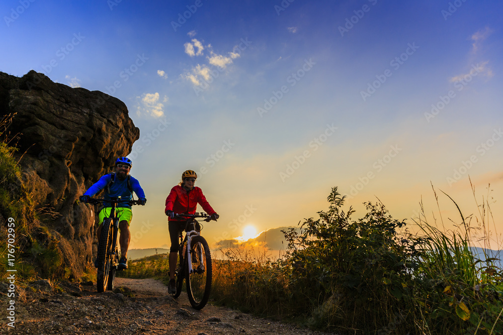Fototapeta premium Cycling women and man riding on bikes at sunset mountains forest landscape. Couple cycling MTB enduro flow trail track. Outdoor sport activity.