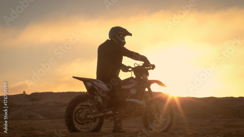 Professional FMX Motorcycle Rider Rests on His Bike and Overlooks Hard Sandy Off-Road Terrain. Sun is Setting.